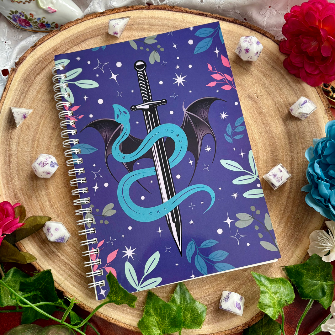Notebook with dragon and sword design on a decorative surface with flowers and dice.