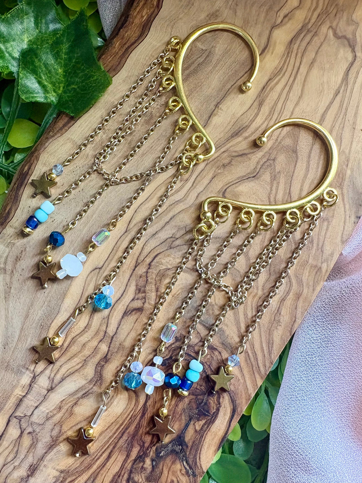 A pair of gold-toned huggie hoop earrings with dangling chains, star charms, and colorful beads, displayed on a textured wooden surface with green leaves and a pink fabric accent.