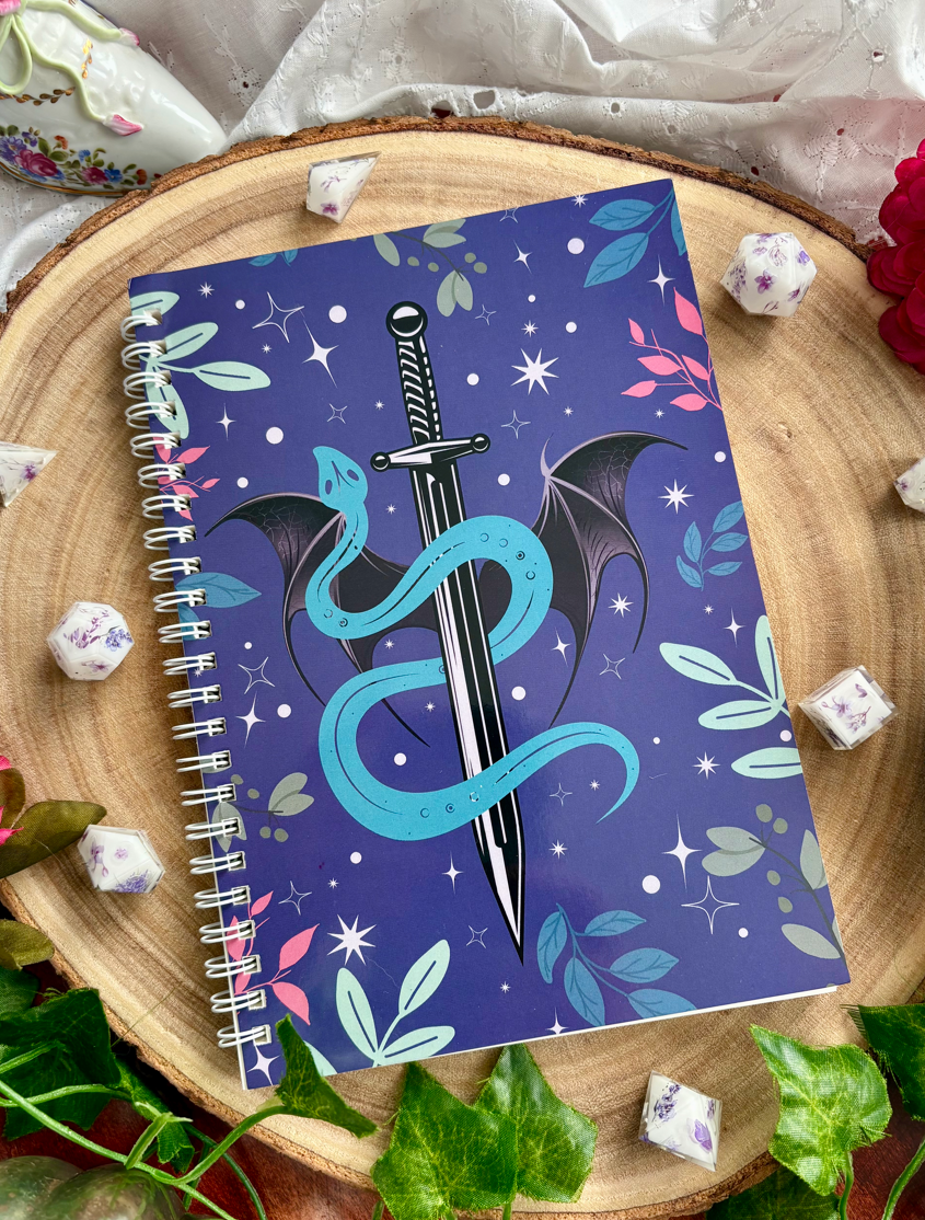 Notebook with a dragon and sword design on a decorative surface with flowers and leaves.