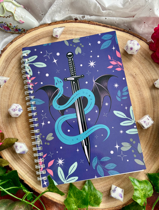 Notebook with a dragon and sword design on a decorative surface with flowers and leaves.