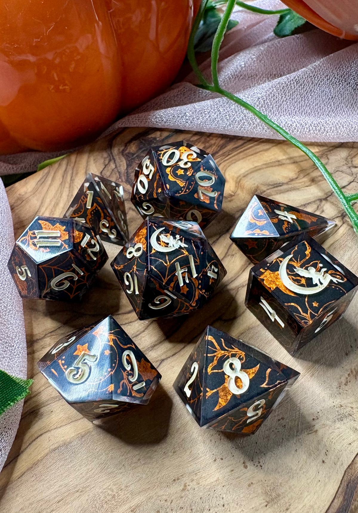 A set of black polyhedral tabletop gaming dice with orange and white Halloween-themed graphics, arranged on a wooden surface with pumpkins and fabric in the background.