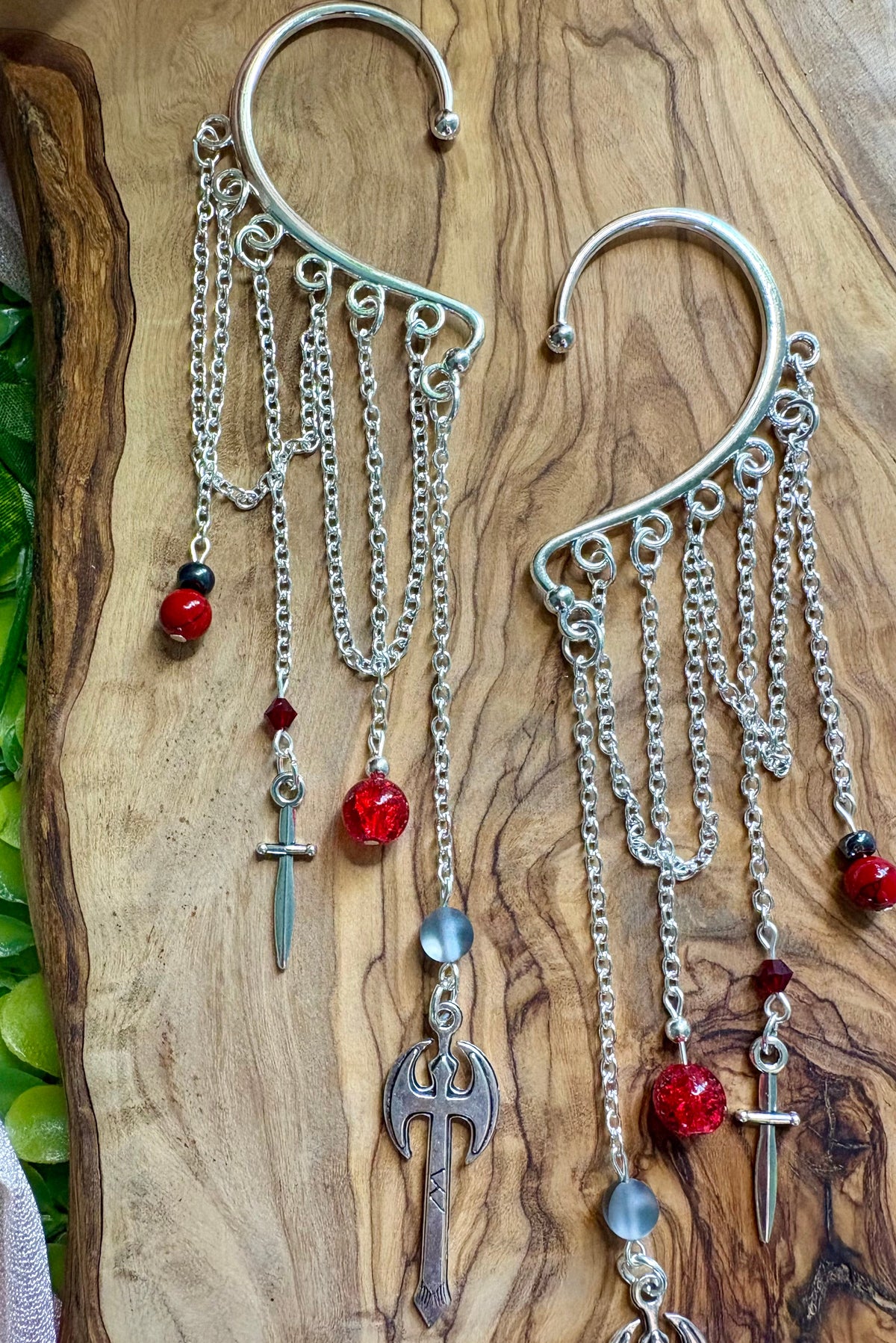 A pair of silver-toned dangle earrings with chains, red beads, small sword and axe charms, displayed on a wooden background/