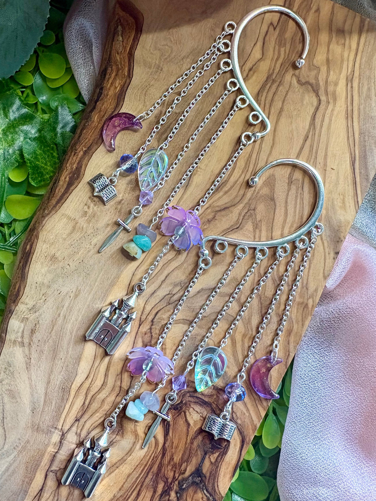 A pair of silver-toned dangle earrings with charms including castles, crescent moons, and iridescent beads, displayed on a wooden board with green leaves and a pink cloth.